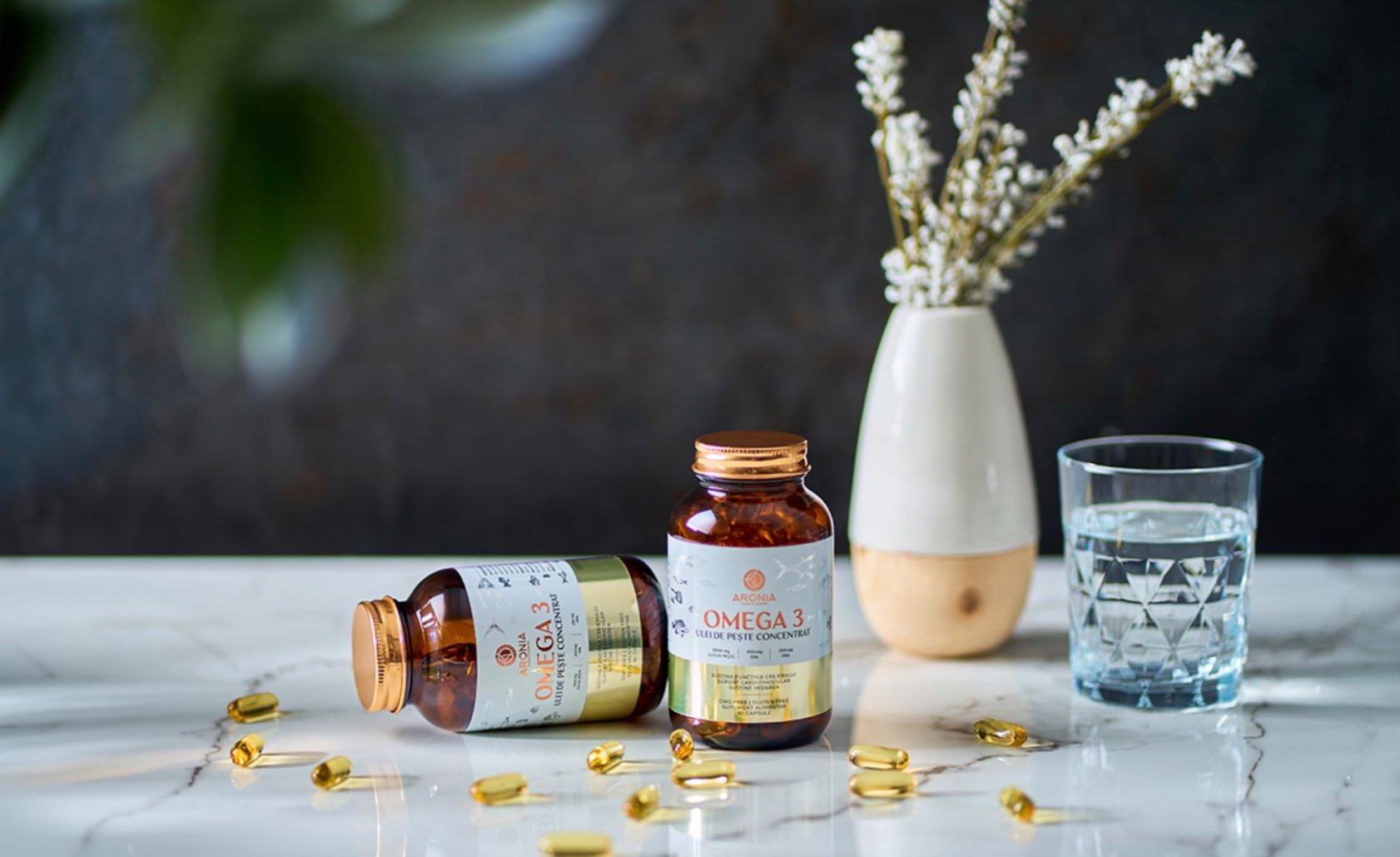 Care sunt cele mai recomandate suplimente alimentare (suplimente cu Omega 3, sticlute maro pe un blat de marmura alba, capsule galbene, vaza cu flori albe)